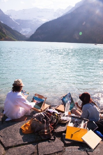 Two painters sit on the steps on the edge of Lake Louise with their equipment spread out around them.