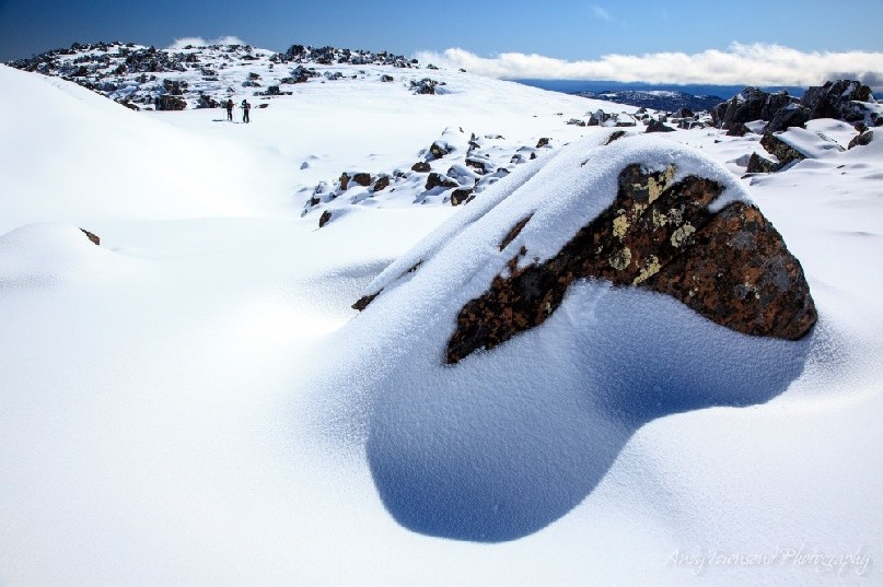 A large snow-covered boulder casts a shadow at the top of the Rodway range, with two backcountry skiers in the distance.