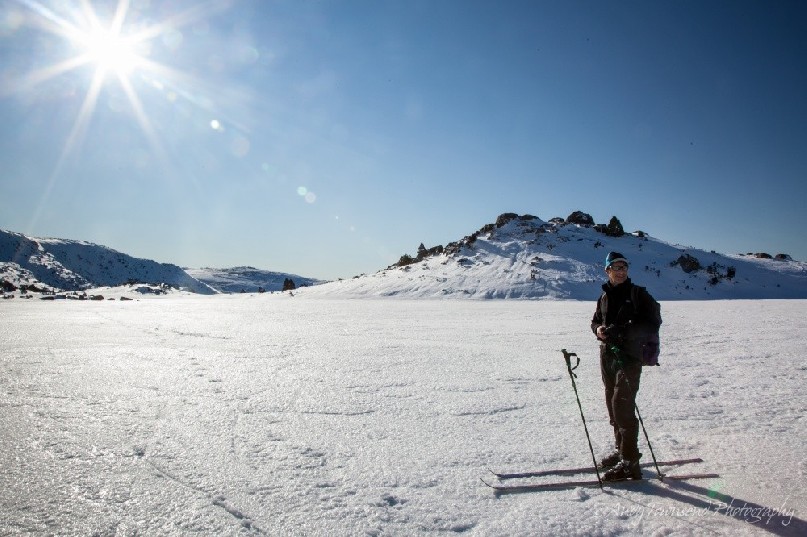 A cross-country skier pauses on the frozen Johnston tarn with a clear blue sky and bright sun behind.