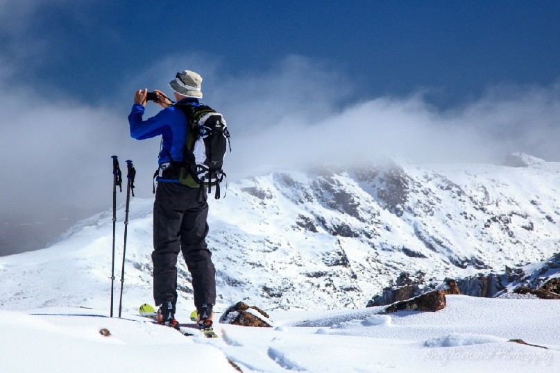 A skier pauses to photograph the mountain vista with a cloud capped mountain in the background.