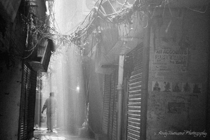 Rays of sunlight shine through dust and chaotic electrical wiring as a person walks along a narrow alleyway.