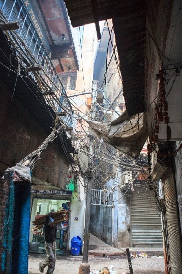 A man balancing wood over his shoulder walks into alley with a view above showing chaotic electrical wiring.