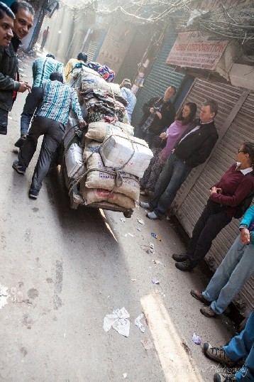 A group of men pushing a laden cart through a narrow alleyway watched on by a group of tourists.