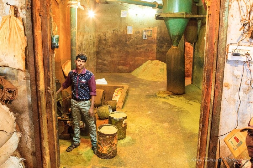 A worker operates the controls of a large spice grinder. A bag of ground coriander fills in the background.