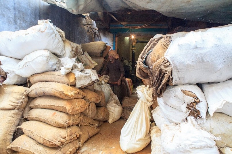 A spice worker makes his way along large bags of spice stacked under a small tarpaulin.