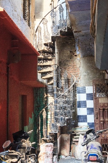 A wrought iron spiral staircase makes its way out of a dusty lane filled with old tiles and motorbikes.