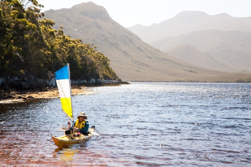 A sea kayaker under sail comes into land at a beach in Bathurst Harbour, Southwest Tasmania with peaks in the distance.