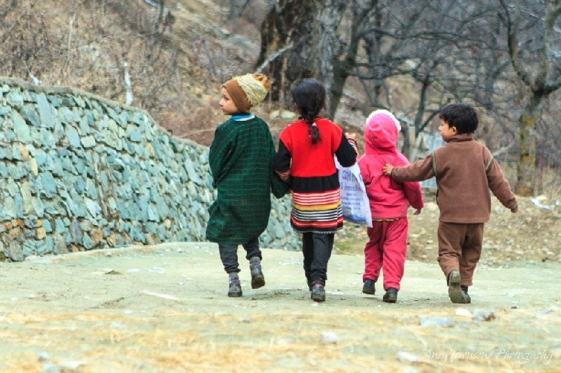 A group of four young children walk together, supportively, down a dirt path near Targmarg village.
