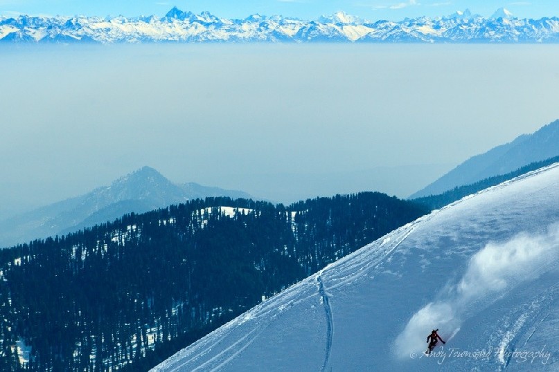 A snowboarder makes their way down an open ridgeline with the distant Himalayan mountains breaking through a layer of fog.