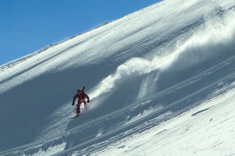 A plume of snow behind a snowboarder in the sunshine with blue sky.