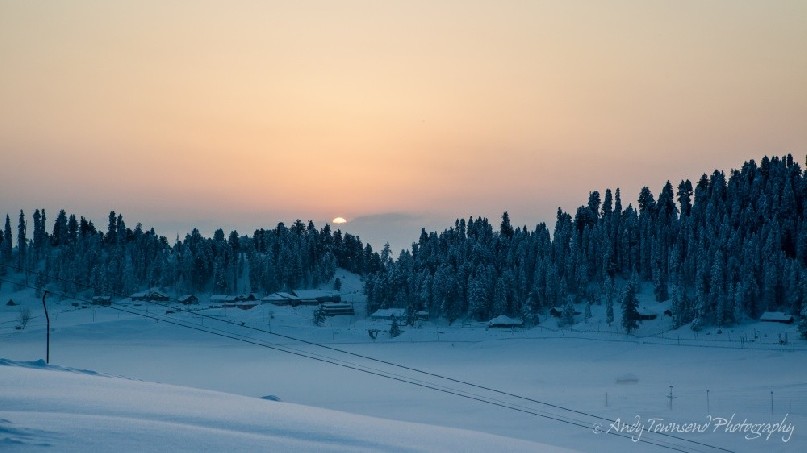 Sunrise breaking through a cloud band over a ridge of trees with fresh snow.