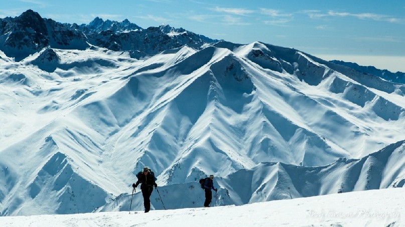 Two skiers skinning up towards the summit of Mt Apharwat with distant mountain range behind.