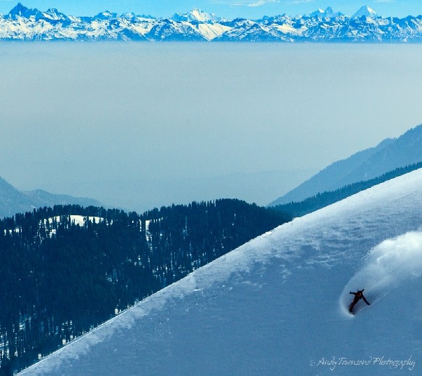 A snowboarder with plume of snow behind arcs down a slope with the distant Himalayan mountains behind.