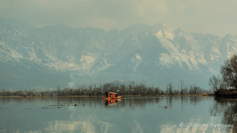 A distant shikara reflects in the stillness of Dal Lake with snow-covered mountains in the distance