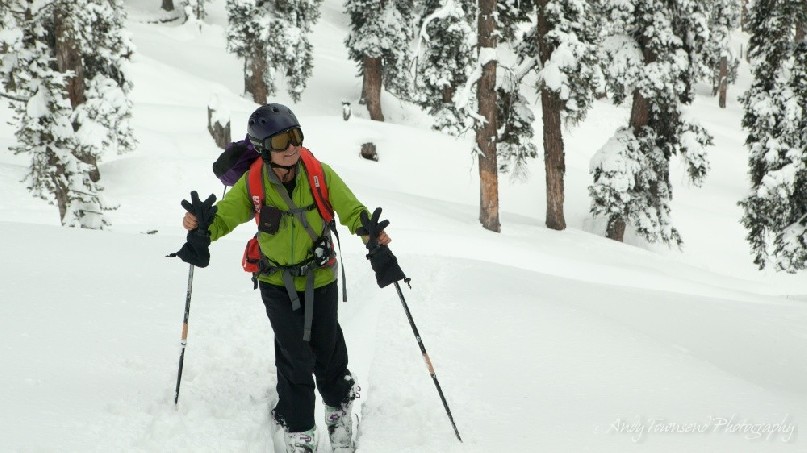A smiling female skier skins up through a snow-covered forest.
