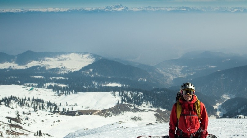A female snowboarder pauses atop a ridge with distant fog in the valley behind and mountains of the Himalaya above.
