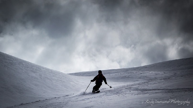 A female telemark skier in mid-turn makes their way down an open slope with dark clouds above.