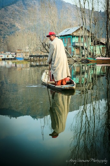 A man standing in a wooden boat wearing traditional phiren is reflected into the still waters of Dal Lake.