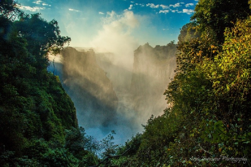 Sunlight on the vegetated cliffs surrounding Victoria Falls with mist from the falls rising in the distance.